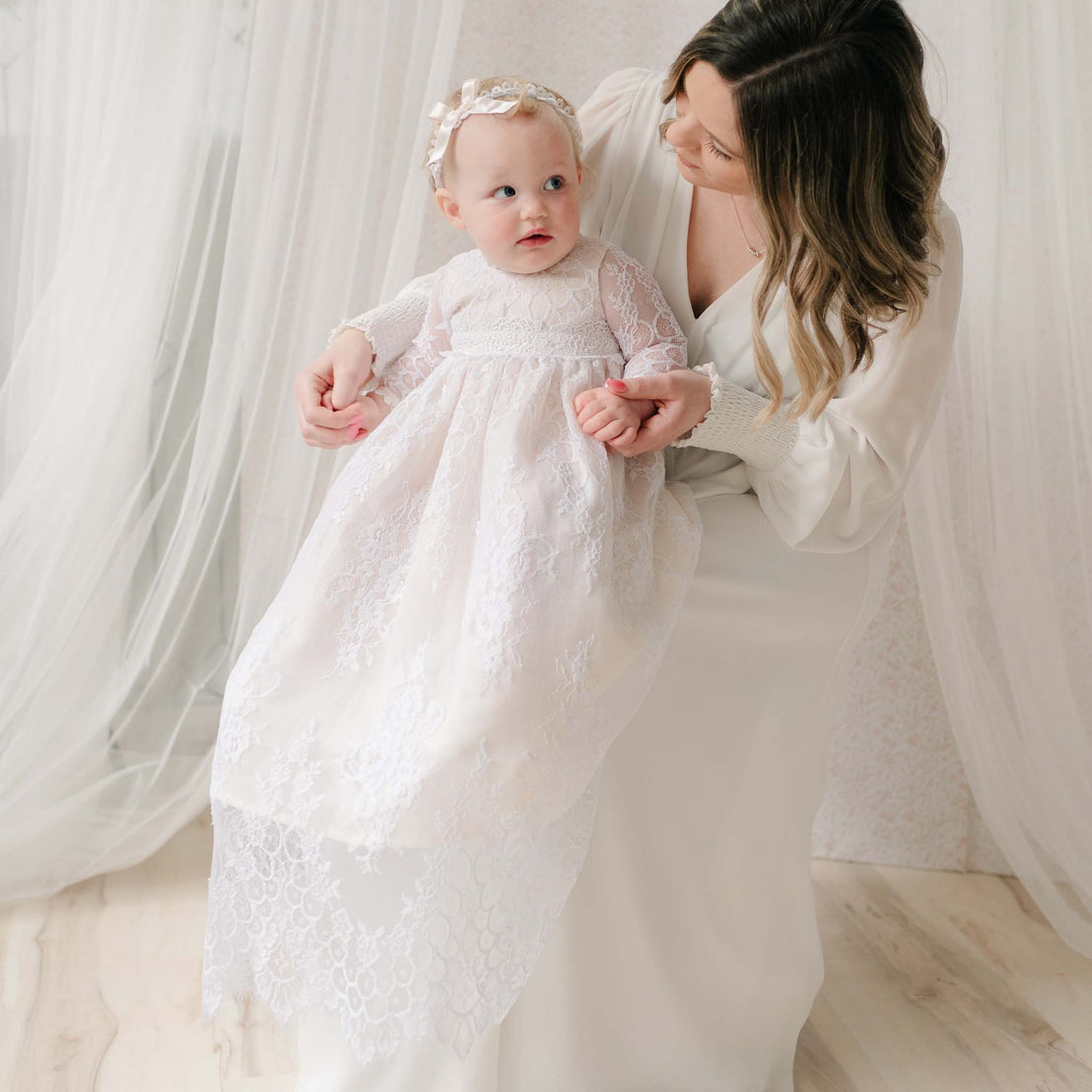 Woman holding a baby dressed in a white lace dress and headband against a soft, neutral background