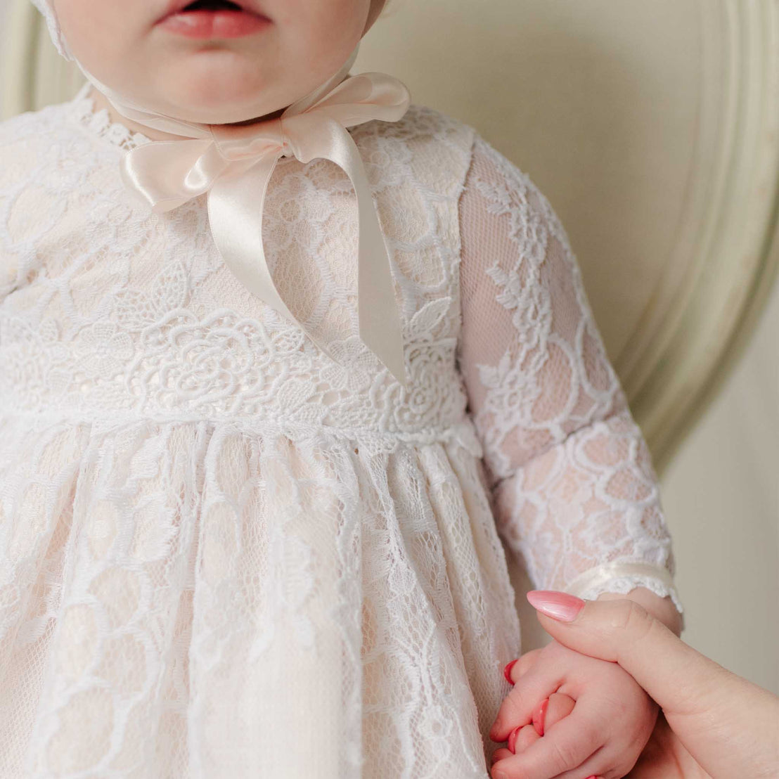 Close-up of a baby wearing a white lace dress with a bow, holding an adult's hand.