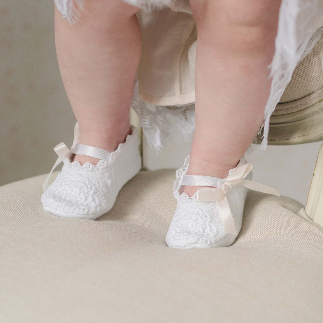 Baby's feet wearing white lace shoes with pink ribbons on a neutral background
