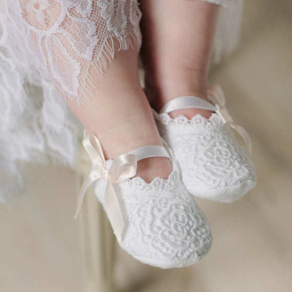 White lace baby shoes with pink ribbons worn by a child in a white dress.