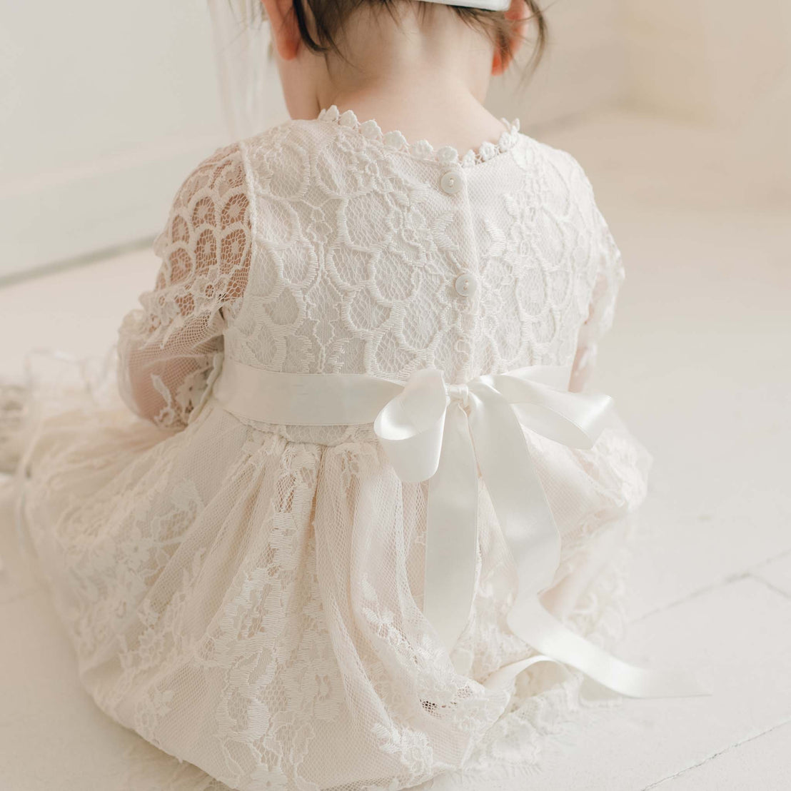 A toddler with dark hair is seated on the floor, facing away from the camera. The child is wearing a Juliette Romper Dress, an intricate white lace outfit featuring a floral pattern and a silk ribbon tie in a bow at the back, adorned with pearl-like buttons. The setting appears light and airy.