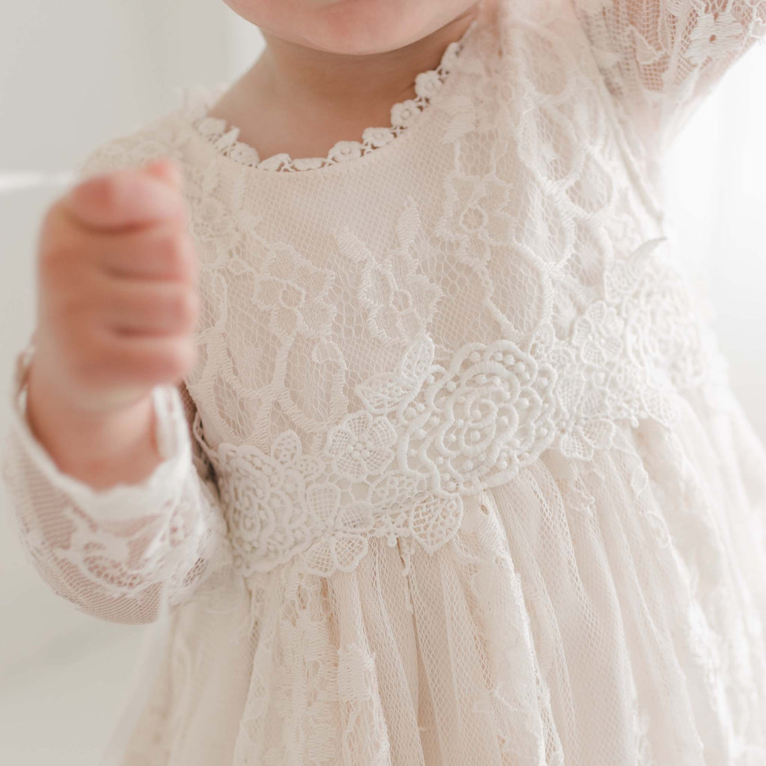 Close-up of a young child wearing the Juliette Romper Dress, a delicate white lace garment adorned with an intricate floral pattern and a beautifully detailed waistband. The dress features a silk ribbon tie at the back, adding to its charm. The child's upper body and part of their face are visible as they reach out with both hands.