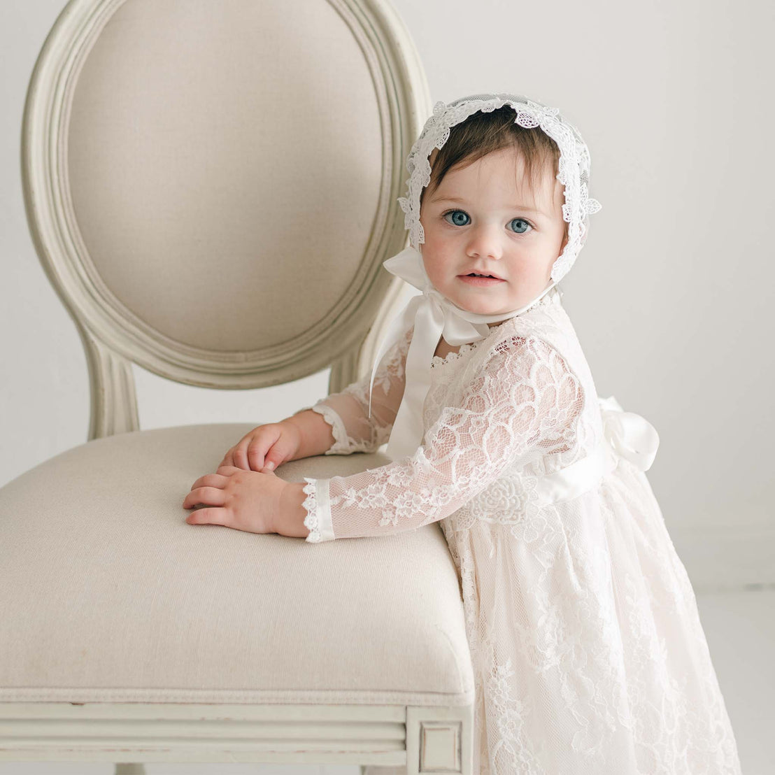 A young child with dark hair, wearing a white lace bonnet and the Juliette Romper Dress, which is adorned with floral pattern lace, stands next to a beige chair with curved wooden frame and upholstery. The blue-eyed child is looking directly at the camera. The background is white and minimalistic.