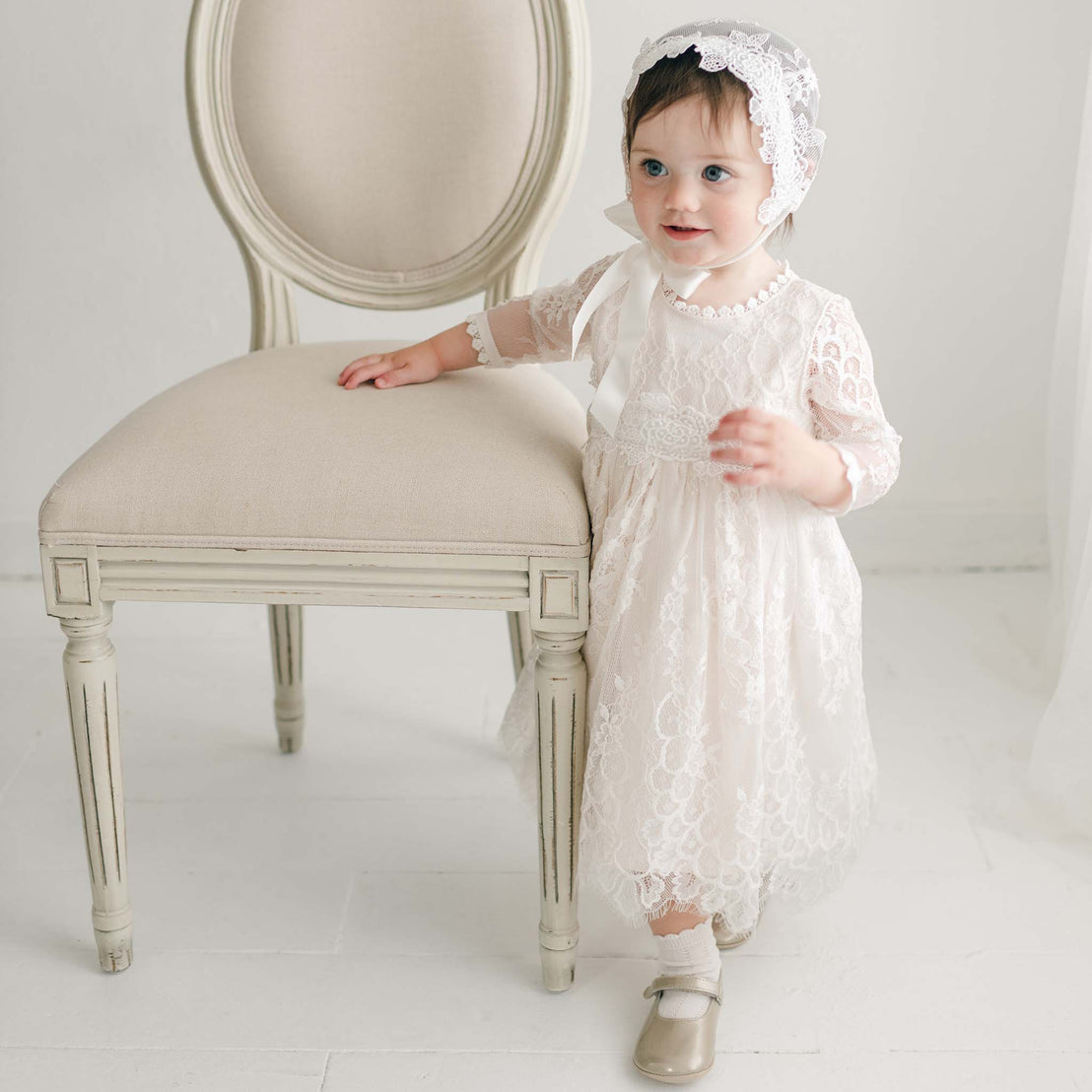 A toddler stands next to a beige, upholstered chair. She is wearing the Juliette Romper Dress, a delicate white lace dress with a floral pattern and a matching lace bonnet. She looks slightly to the side, holding the chair with one hand while her other hand rests by her side.