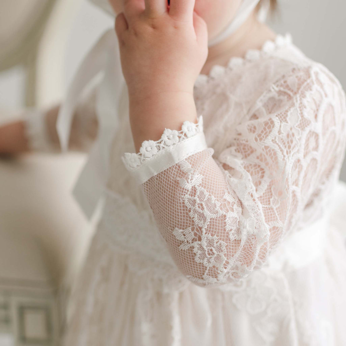 A close-up of a young child dressed in the delicate Juliette Romper Dress with long sleeves. The child’s hand is raised to their mouth, showcasing the silk ribbon tie around one wrist. The soft texture and intricate floral pattern of the lace are emphasized in the image.