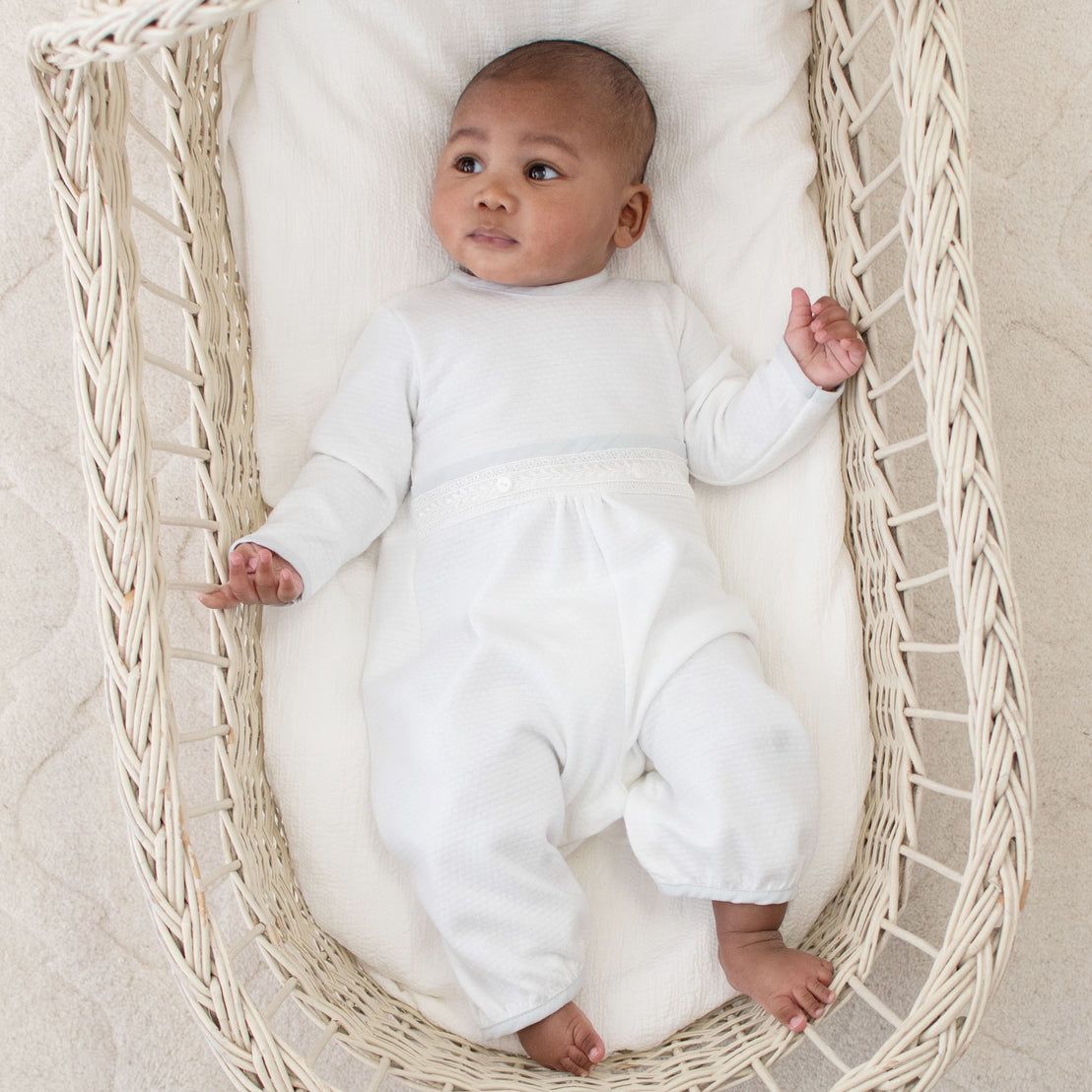 A baby laying in a crib and wearing the Harrison Long Sleeve Romper made from textured white cotton and featuring ivory Venice lace and a blue cotton/linen edging