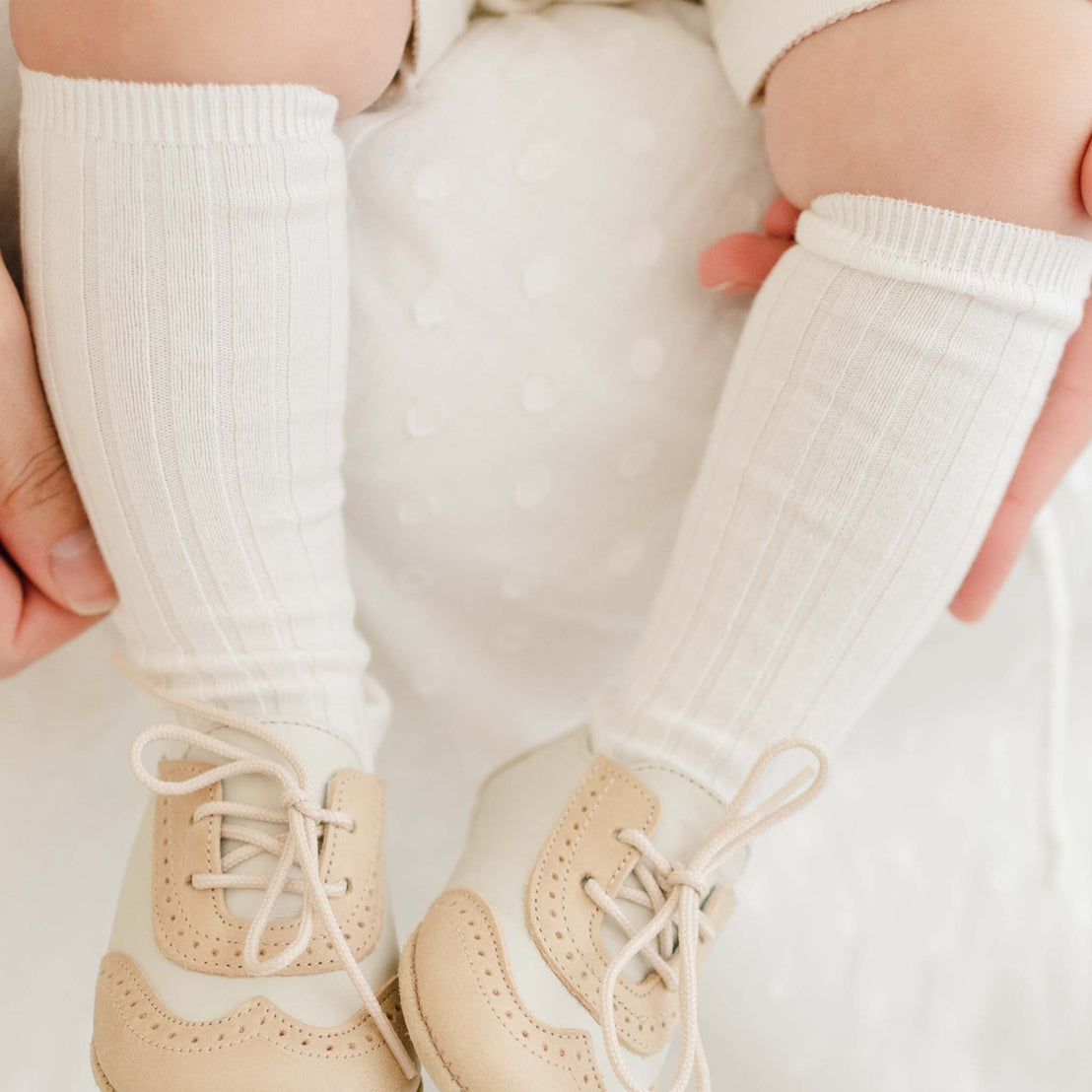 Close-up image of a baby's feet in Cream Ribbed Knee Socks, set against a vintage, white dotted fabric background. Suitable for baptism or christening events.