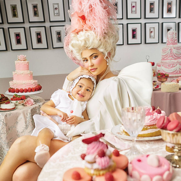 A woman with elaborate hair and white dress holds a child on her lap in a room filled with pink and white cakes and desserts. The walls display framed pictures. The theme is elegant and whimsical, resembling a Marie Antoinette style setting.