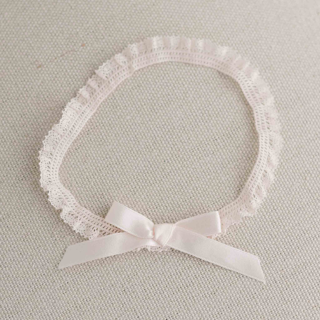 A delicate Ava Headband made from a blush pink lace with a matching silk ribbon bow, placed on a light grey textured background.