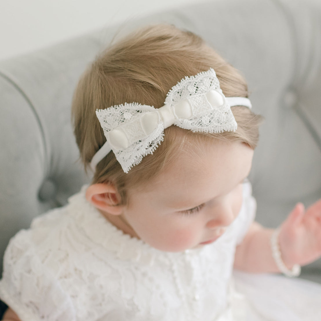 A baby with light hair wearing Aria Christening Gown and an Aria Lace Bow Headband, looking downward, seated on a gray upholstered chair.