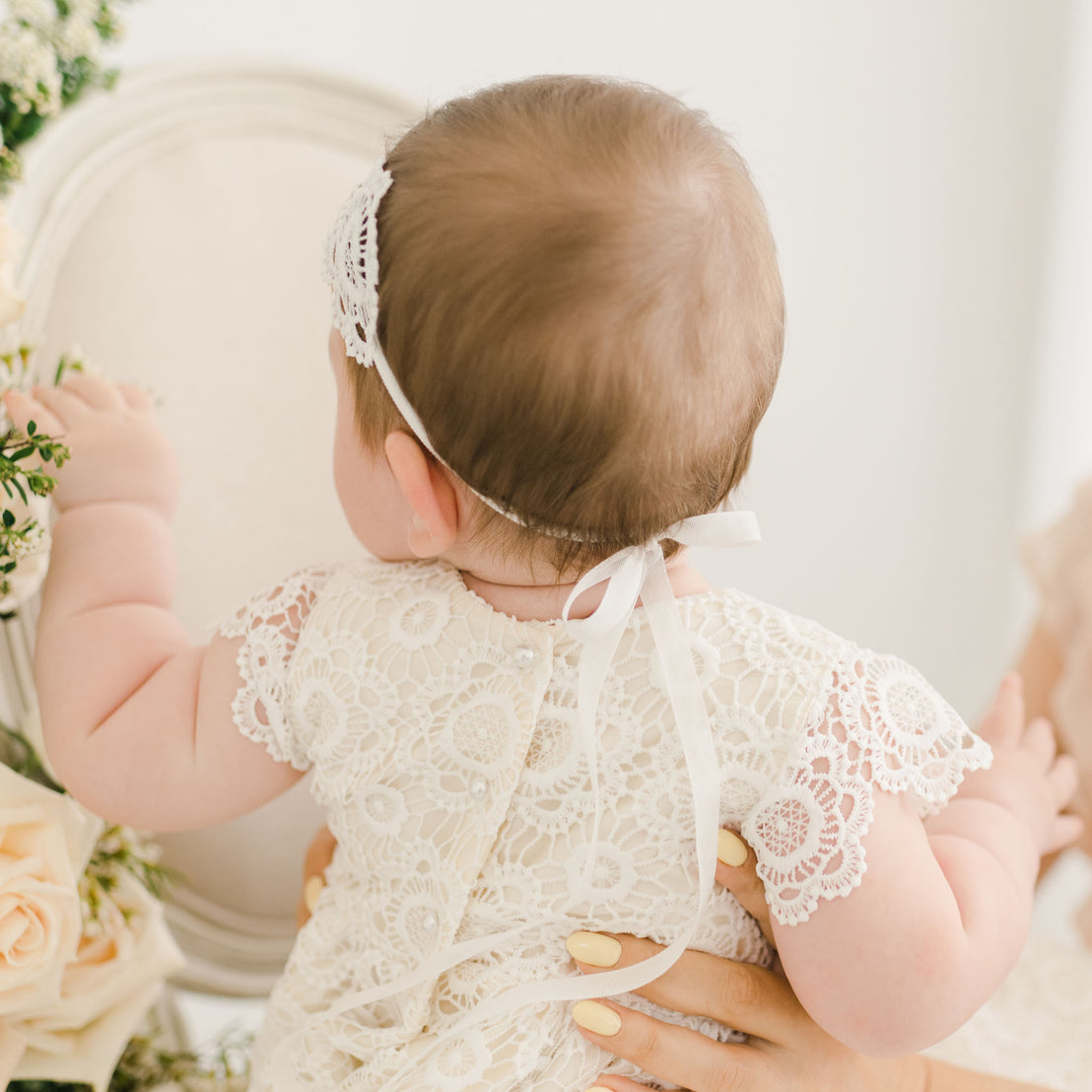Back view of a baby wearing the Poppy Lace Headband with a white ribbon tied at the back, paired with the matching dress adorned with pearl button details.