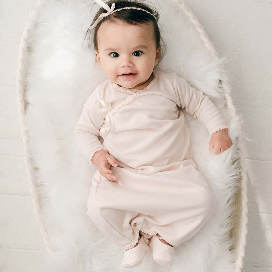 A baby wearing the wearing the Ava Newborn Layette & Headband, a pima cotton layette in a shade of blush pink and matching blush ribbon bow headband, lies smiling in a white fluffy blanket.