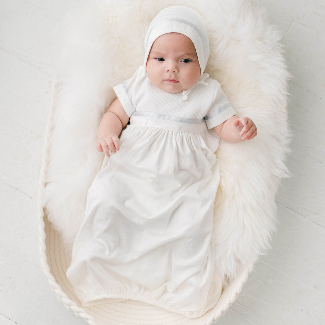 Newborn baby boy sitting in a white fur-lined crib. He is wearing the Owen Layette and Quilted Newborn Bonnet.