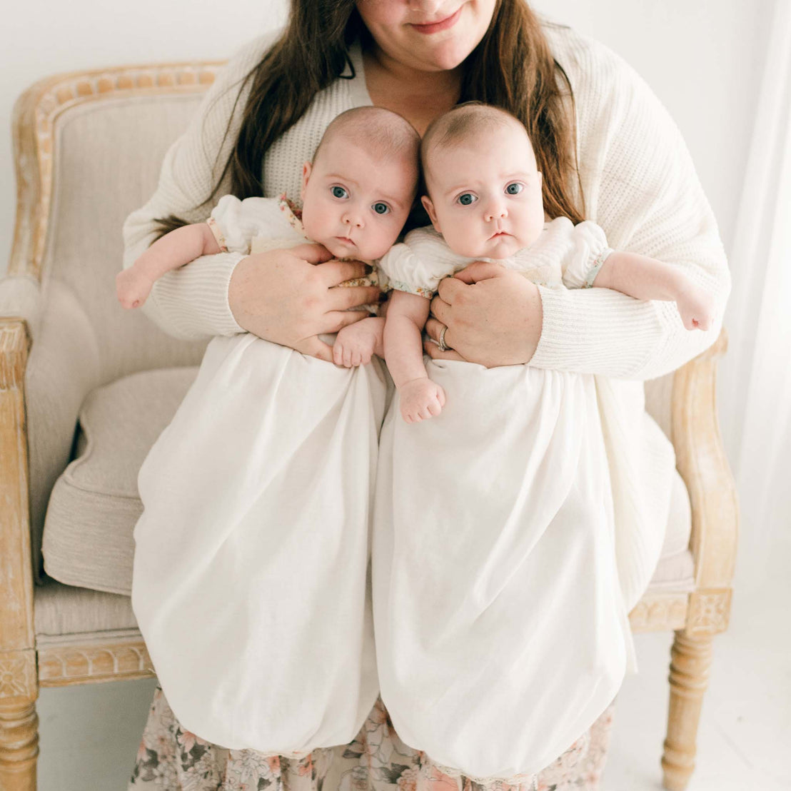 Two twin baby girls, both being held by their mother, and wearing the Eloise Layette Gown.
