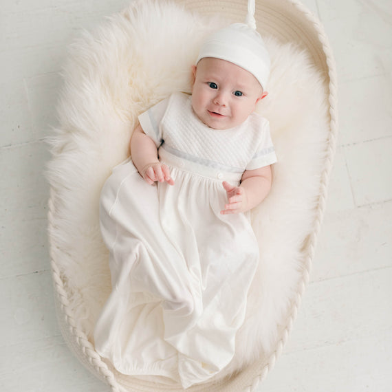 Baby boy sitting in a white fur-lined crib. He is wearing the Owen Layette and Knot Cap