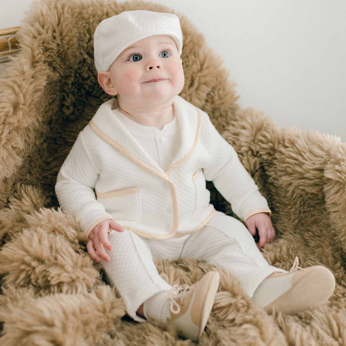 Baby sitting on a chair with brown fur. He is wearing the Liam 3-Piece Suit made with soft ivory quilted cotton and champagne silk trim. Underneath he is wearing a cotton onesie