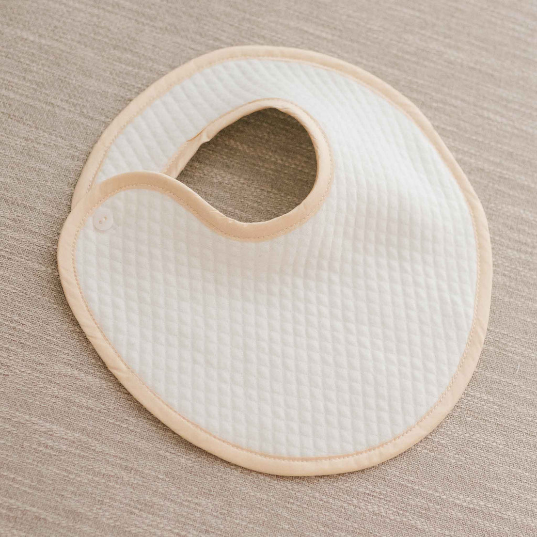 Flat lay photo of Liam Boys Bib. It is crafted with soft quilted cotton in ivory and is detailed with a silk champagne trim