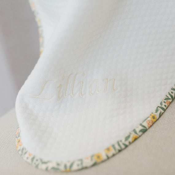 Close up flat lay photo of the Lillian Personalized Blanket on a beige background. Details show yellow floral trim and the name "Lillian" embroidered on the corner of the blanket.
