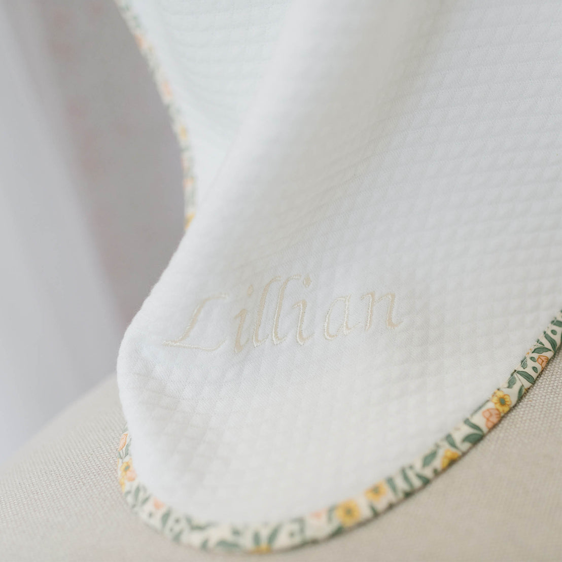 Close up flat lay photo of the Lillian Personalized Blanket on a beige background. Details show yellow floral trim and the name "Lillian" embroidered on the corner of the blanket. Perfect for baptism gift from godparents.