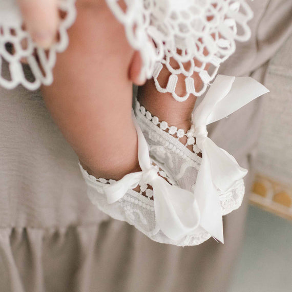 Baby wearing Lily booties that are made of soft cotton lace in light ivory with silk ribbon ties