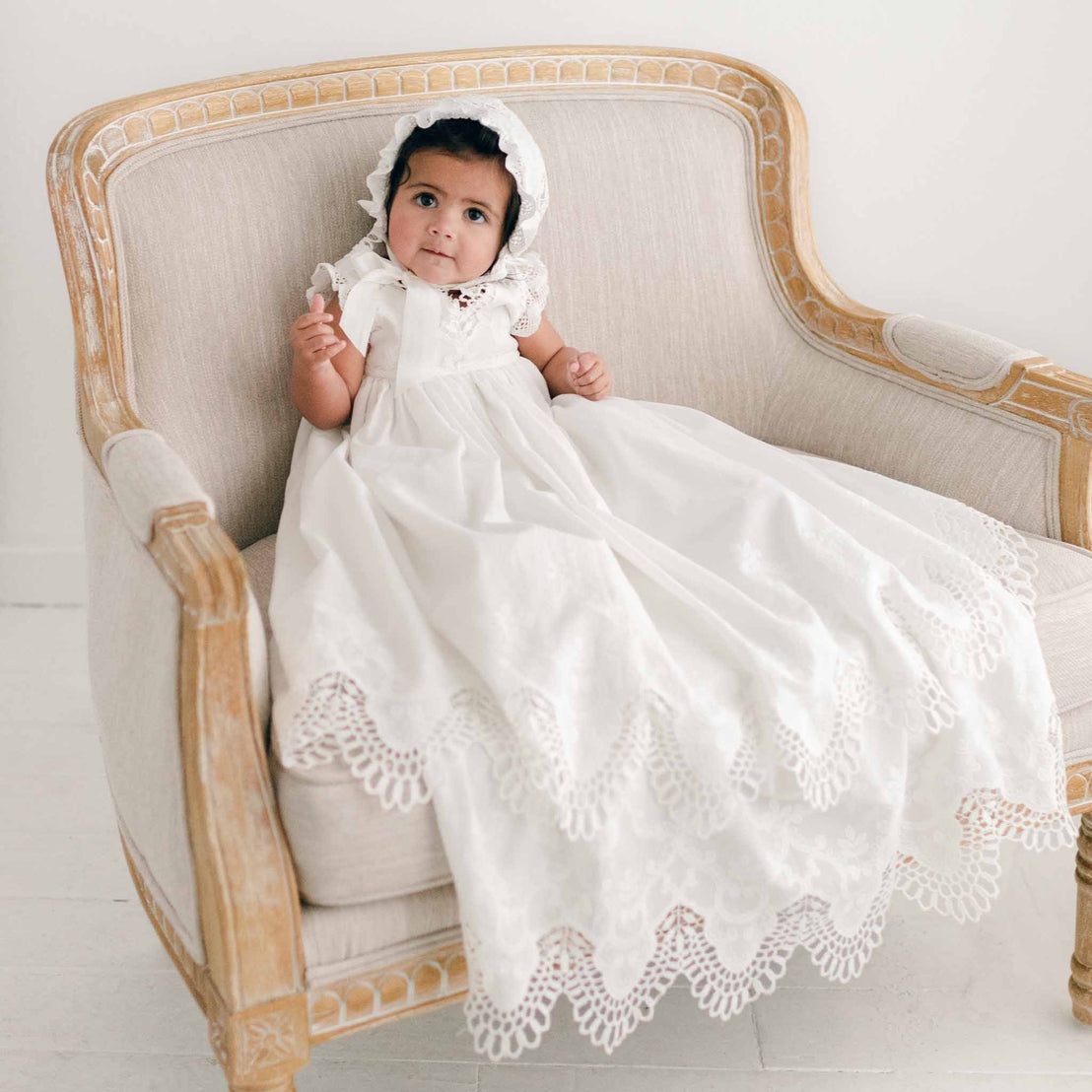 Photo of a baby sitting on a chair wearing the Lily Christening Gown and Bonnet featuring an elegantly detailed cotton eyelet lace with a scalloped edge