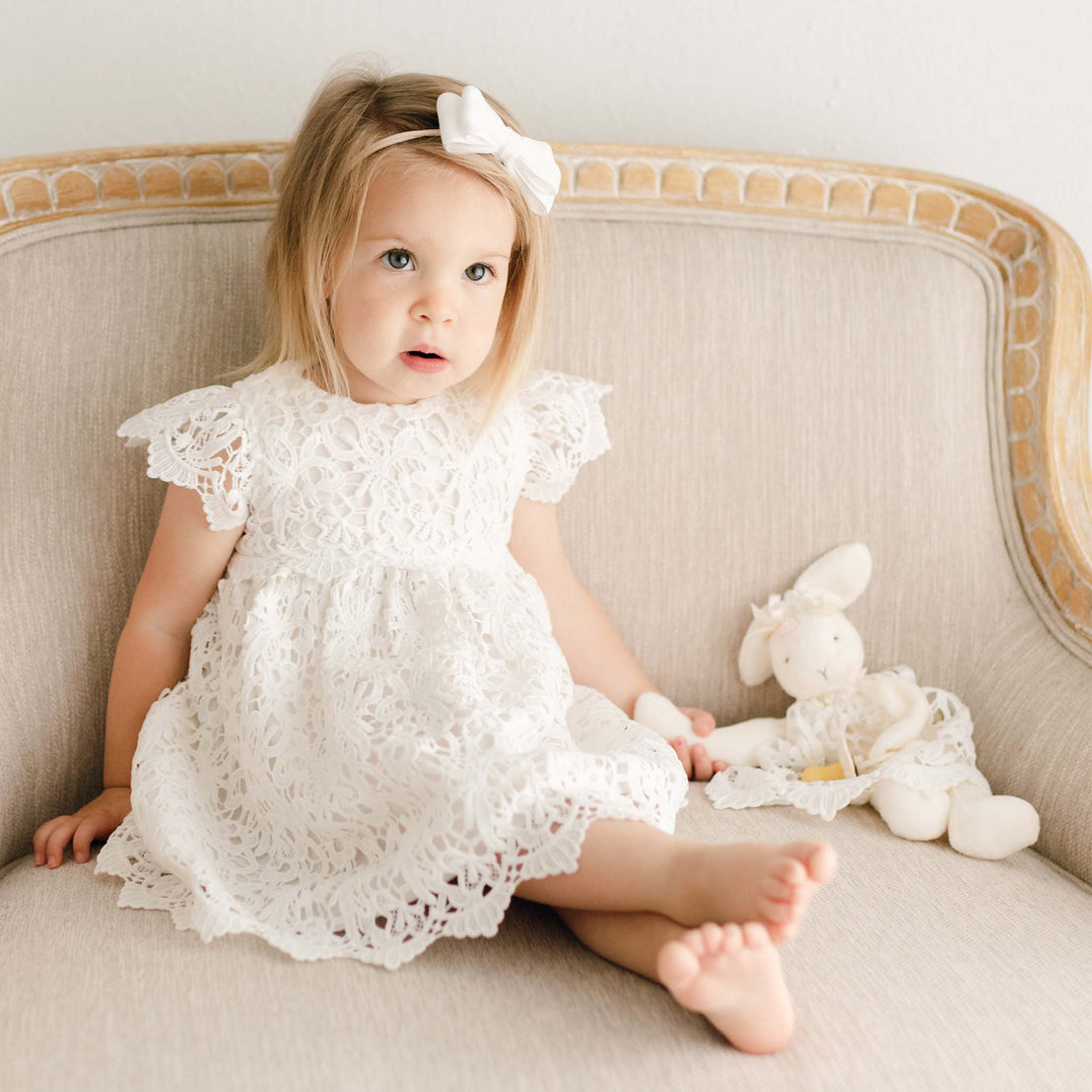 Baby girl wearing the Lola Dress and Lola velvet headband. Dress is made with cotton lining in light ivory featuring an all-over lace overlay. She is holding a Lola Silly Bunny Buddy pacifier holder. 