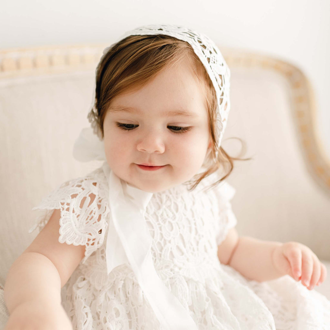 Baby girl on a couch and wearing the Lola Christening Gown and Bonnet, a baptismal gown made with a cotton undergown in light ivory featuring an all-over lace overlay.