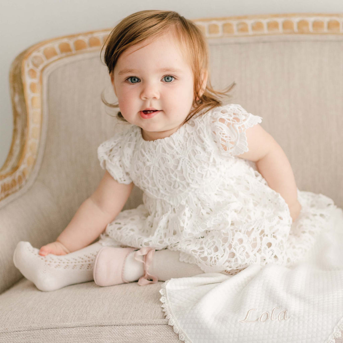 Baby girl sitting on a couch and wearing the Lola Dress. The dress is made with a cotton lining in light ivory featuring an all-over lace overlay.