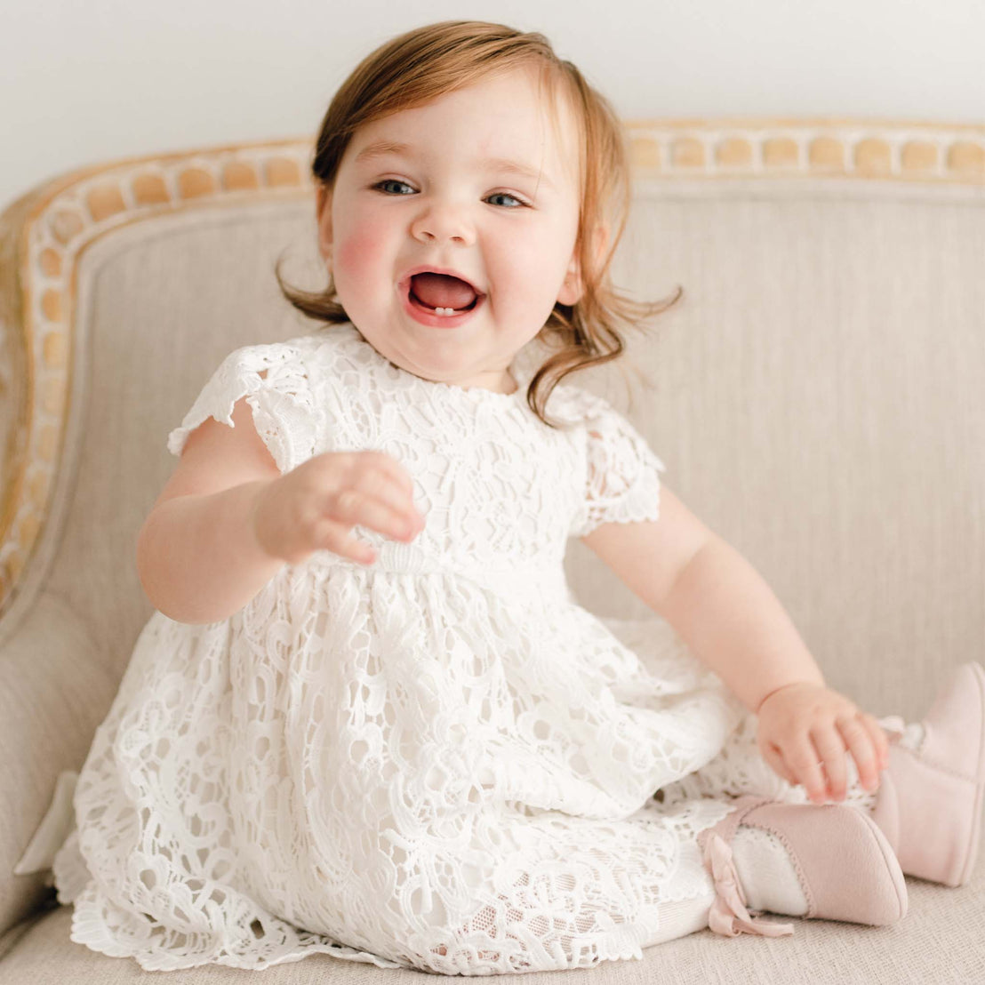 Smiling baby girl wearing the Lola Dress. Dress is made with cotton lining in light ivory featuring an all-over lace overlay.