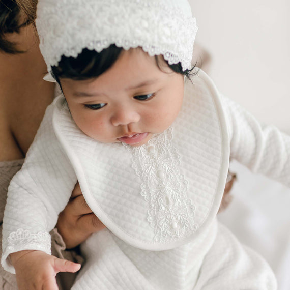 A baby girl wearing a Madeline Bib over the Madeline Quilted Newborn Romper and Bonnet. She is being held by her mother and looking to the side with a thoughtful expression.