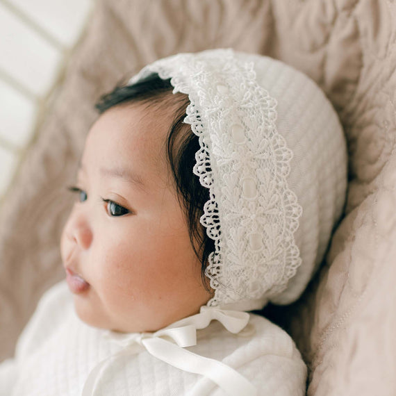 A baby wearing a Madeline Quilted Newborn Bonnet lies on a quilted beige blanket. The baby has chubby cheeks and is dressed in a white outfit, looking off to the side. The soft lighting gives the scene a warm, gentle feel.