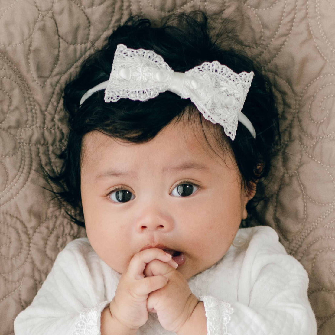 A baby with curly dark hair lies on a quilted surface, wearing a Madeline Lace Bow Headband. Dressed in a white outfit with lace trim, handmade in the USA, their hands lightly touch their face near their mouth as they gaze directly at the camera.