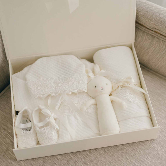 A boutique baby gift box containing a Madeline Newborn Gift Set of heirloom items: a white blanket, a small stuffed toy, baby shoes, and a fabric outfit, all set against a soft, neutral background.