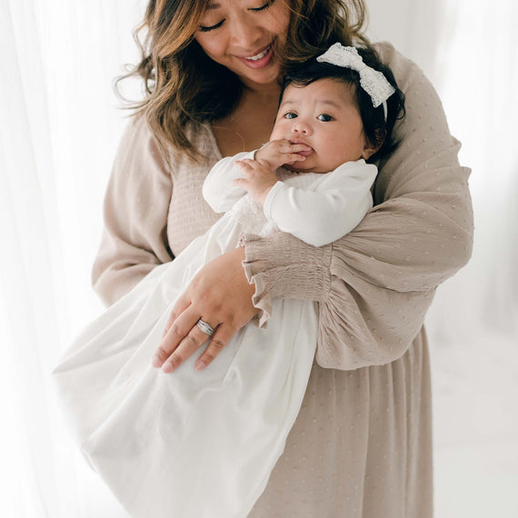 A smiling woman with wavy hair holds a baby dressed in a white pima cotton outfit with a bow headband. The baby, who has a hand in their mouth, is perfectly snug in their Madeline Newborn Gown. The woman wears a light-colored, long-sleeved dress and has a gentle, loving expression as soft, bright light filters in from the background.
