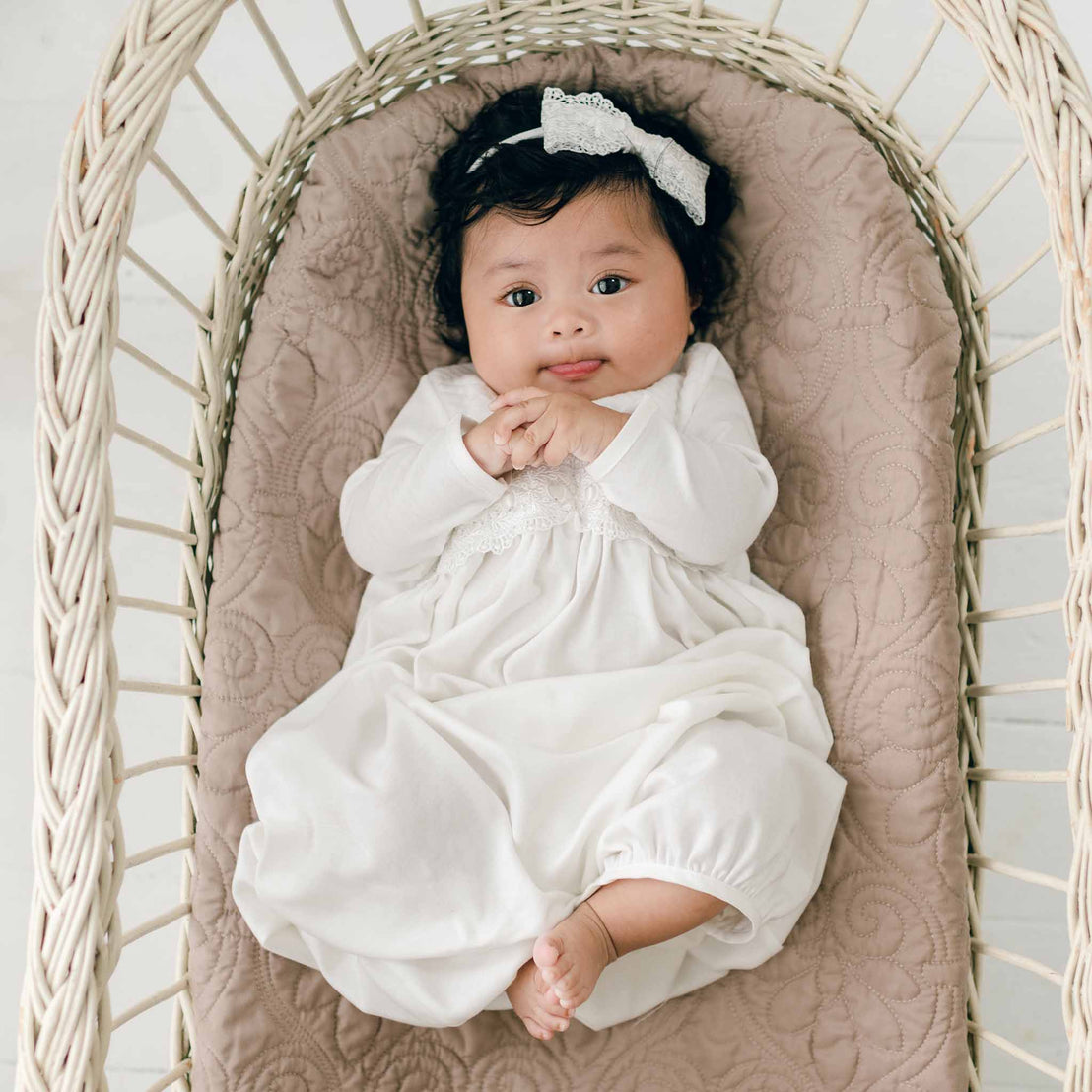 A baby with dark curly hair, wearing a white Madeline Newborn Gown and a white headband with a bow, lies on a quilted beige cushion inside a wicker bassinet. The baby has hands clasped and looks up with a curious expression.