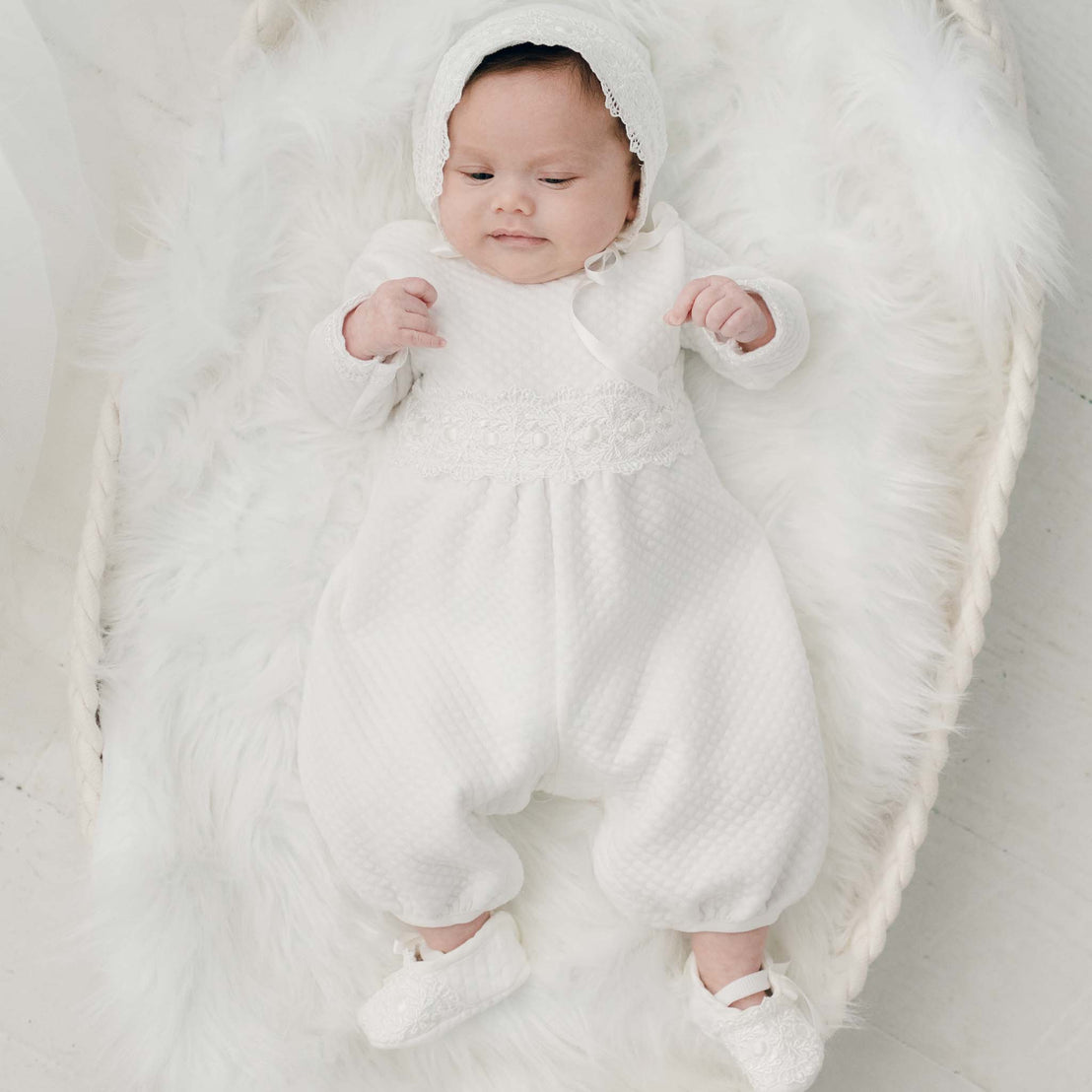 A newborn baby dressed in a Madeline Quilted Newborn Romper with lace details, lying on a soft white fur blanket, gazing gently upward.