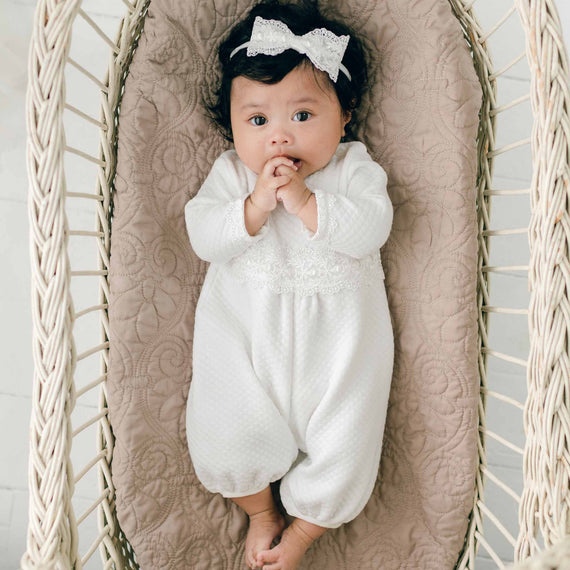 A newborn girl in a Madeline Quilted Newborn Romper and a lace headband lies on a quilted cotton cushion inside a woven basket. The baby has their hands touching their mouth and gazes up, surrounded by softly textured neutral-colored bedding. This adorable scene is handmade in the USA.