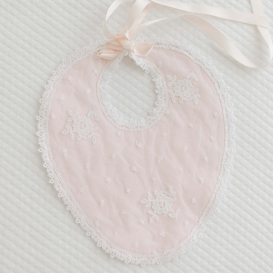 The Melissa Bib is a delicate, pink lace baby bib featuring white floral embroidery and a white ribbon tie. It boasts elegant Venice lace accents and a charming pearl button, with a scalloped edge and small white polka dots, lying flat on a textured white fabric background.