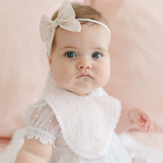 A baby with blue eyes, wearing a white lace dress and the Melissa Bib—a quilted cotton bib adorned with embroidered flowers—gazes at the camera. The baby has a headband featuring a large, light-colored bow and is seated against a soft, peach-colored background.