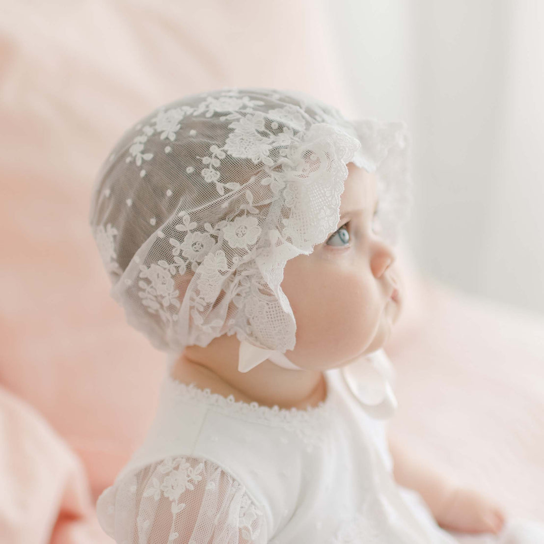 A baby wearing a delicate white Melissa Christening Gown & Bonnet is gazing upwards. The background is softly blurred with pastel pink and white tones, creating a dreamy, serene atmosphere.