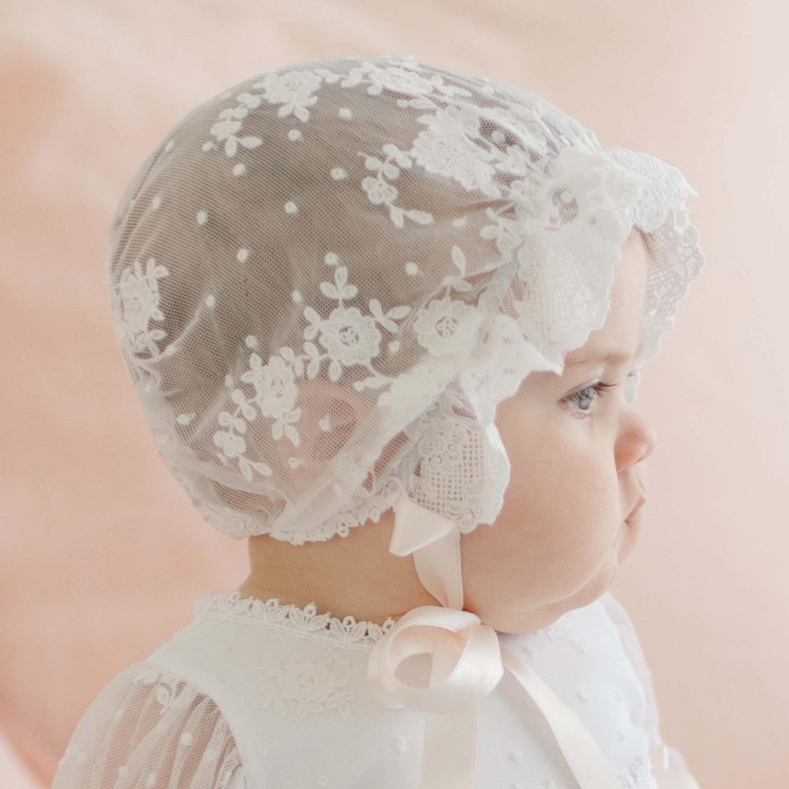 Profile view of a baby girl wearing the Melissa Sheer Bonnet, looking away from the camera. This angle highlights the bonnet’s sheer dotted netting with embroidered floral patterns.