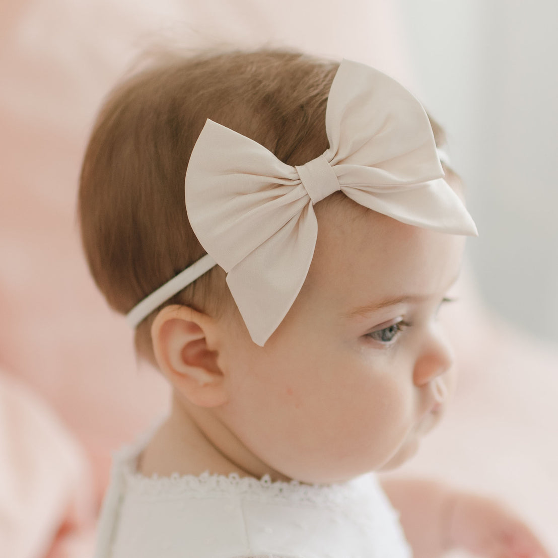 A close-up profile image of a baby. The baby is wearing a large white Melissa Bow Headband and a white outfit with delicate detailing. The background is soft and out of focus, featuring pastel tones. Enhanced with a touch of elegance, the handmade headband adds charm as the baby looks downward slightly, showcasing light brown hair.