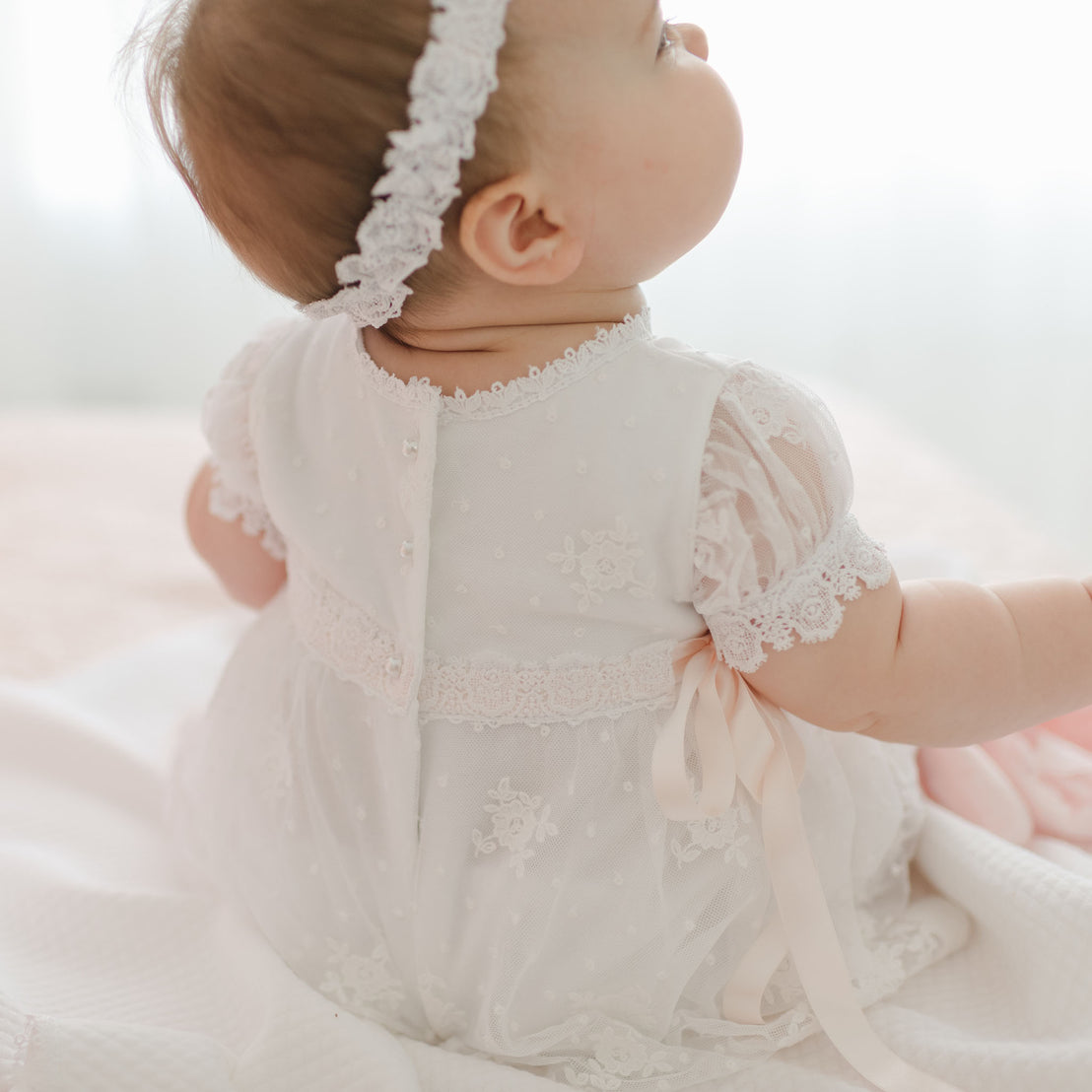 A baby dressed in a white Melissa Christening Gown & Bonnet with Venice lace trim and embroidered details sits on a white blanket. The baby's back is towards the camera, showcasing the delicate lace and buttons on the gown. The scene is softly lit with a blurred background.