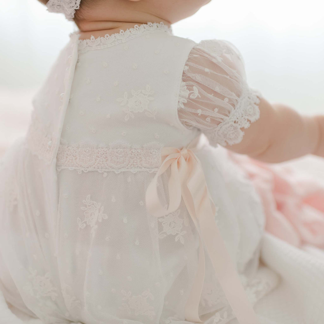 A baby is wearing a delicate, white pima cotton dress with intricate floral embroidery and sheer, patterned sleeves. A soft, pink ribbon bow ties at the back. The baby is sitting and facing away from the camera, showcasing the details of the Melissa Christening Gown & Bonnet adorned with Venice lace trim.