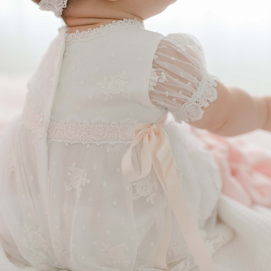 A baby is seen from the back, wearing a delicate Melissa Romper Dress adorned with lace, floral embroidery, and a satin bow. The baby's arms are reaching out, and the soft focus background adds a gentle, serene ambiance to the image.