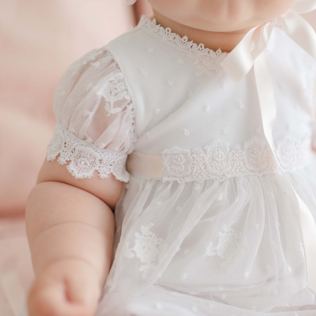 A close-up of a baby wearing a delicate, white Melissa Christening Gown & Bonnet with short, puffed sleeves adorned with intricate Venice lace trim. The baby's arm is extended, and a satin ribbon bow is visible near the neck. The background is softly blurred in pastel tones.