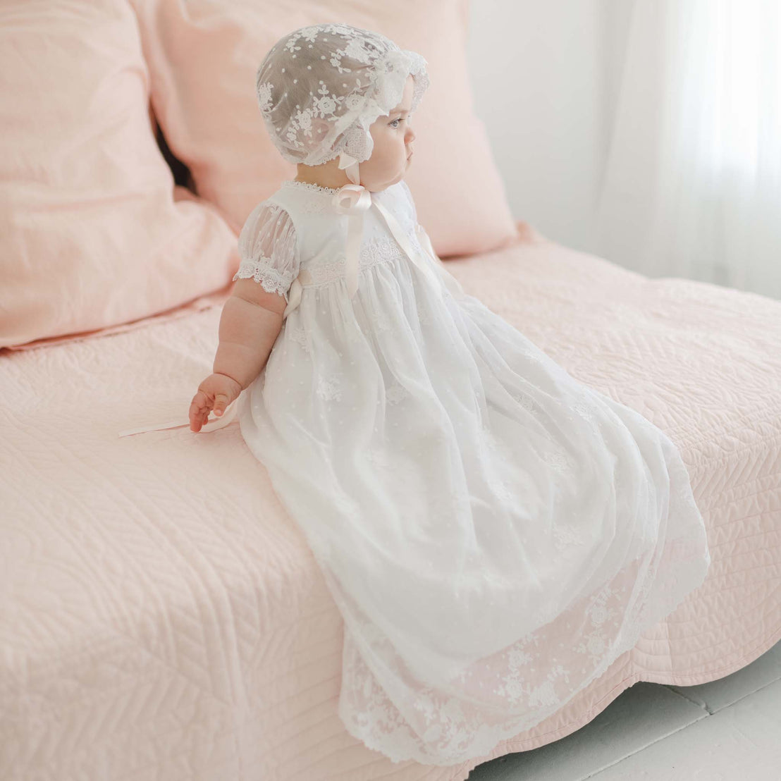 A baby wearing a long, white Melissa Christening Gown & Bonnet sits on a blush pink bed. The baby is turned slightly to the side, looking away. The bed is made with a quilt of the same blush pink color, and the background is softly lit.