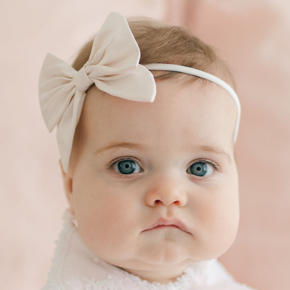 A baby with light skin and blue eyes, wearing a Melissa Bow Headband, gazes directly at the camera. The baby has light brown hair and is dressed in a white outfit. The background is a soft, light pink adorned with delicate baby accessories like handmade headbands and pink silk ribbons.