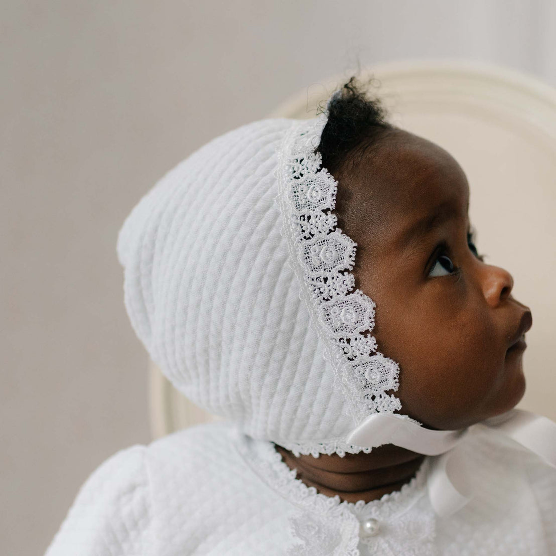Baby wearing a white textured and lace bonnet against a neutral background