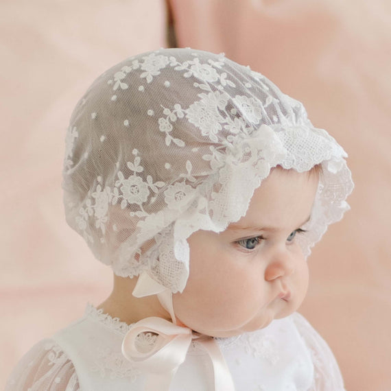 Side profile of a baby girl wearing the Melissa Sheer Bonnet, featuring delicate floral lace and scalloped edges. The bonnet is tied with soft satin ribbons under her chin.